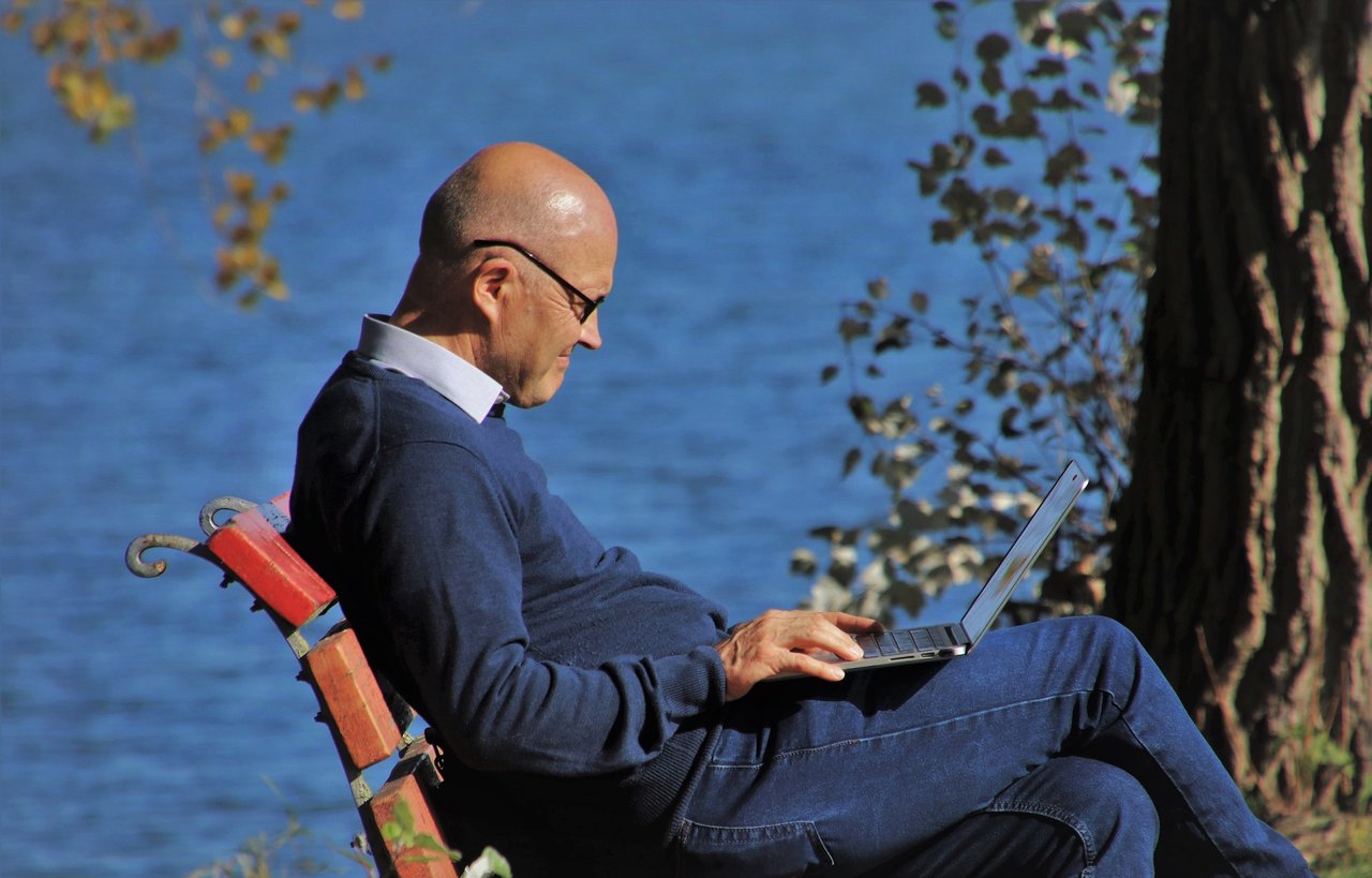 Man sitting on park bench with laptop near the water